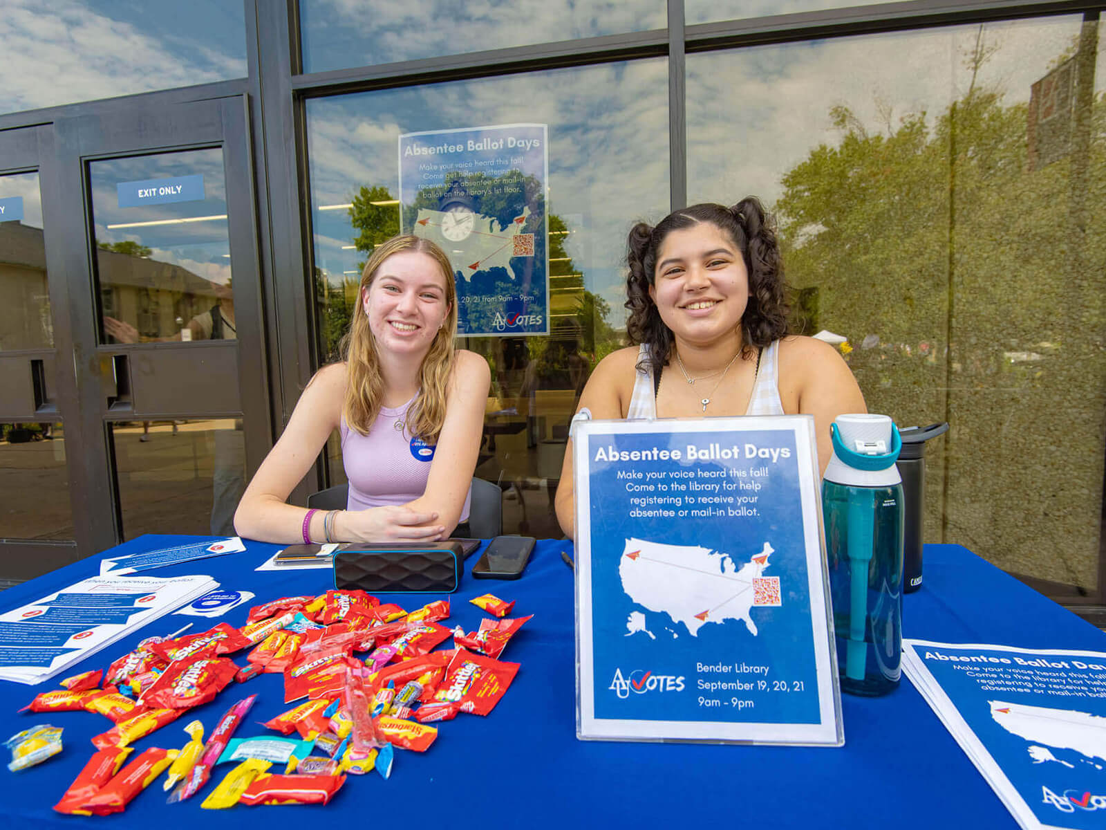 Two women at a table filled with colorful candy, accompanied by a sign, sharing a joyful moment together.