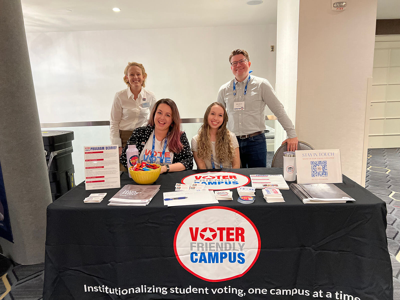A trio of people stands behind a table with a "Voter Camp" sign, encouraging community participation in the voting process.