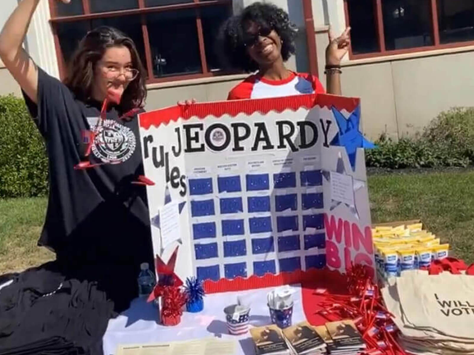 Two women pose together in front of a table featuring a sign that says "Jeoparday.
